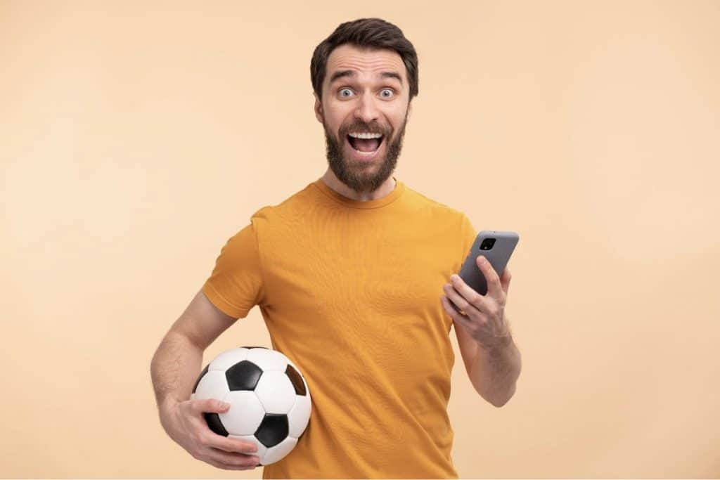 Homem com smartphone em uma mão e uma bola de futebol na outra, animado com apostas esportivas online