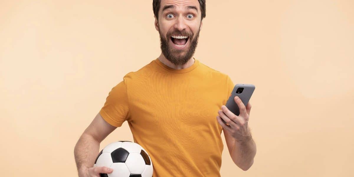 Homem com smartphone em uma mão e uma bola de futebol na outra, animado com apostas esportivas online